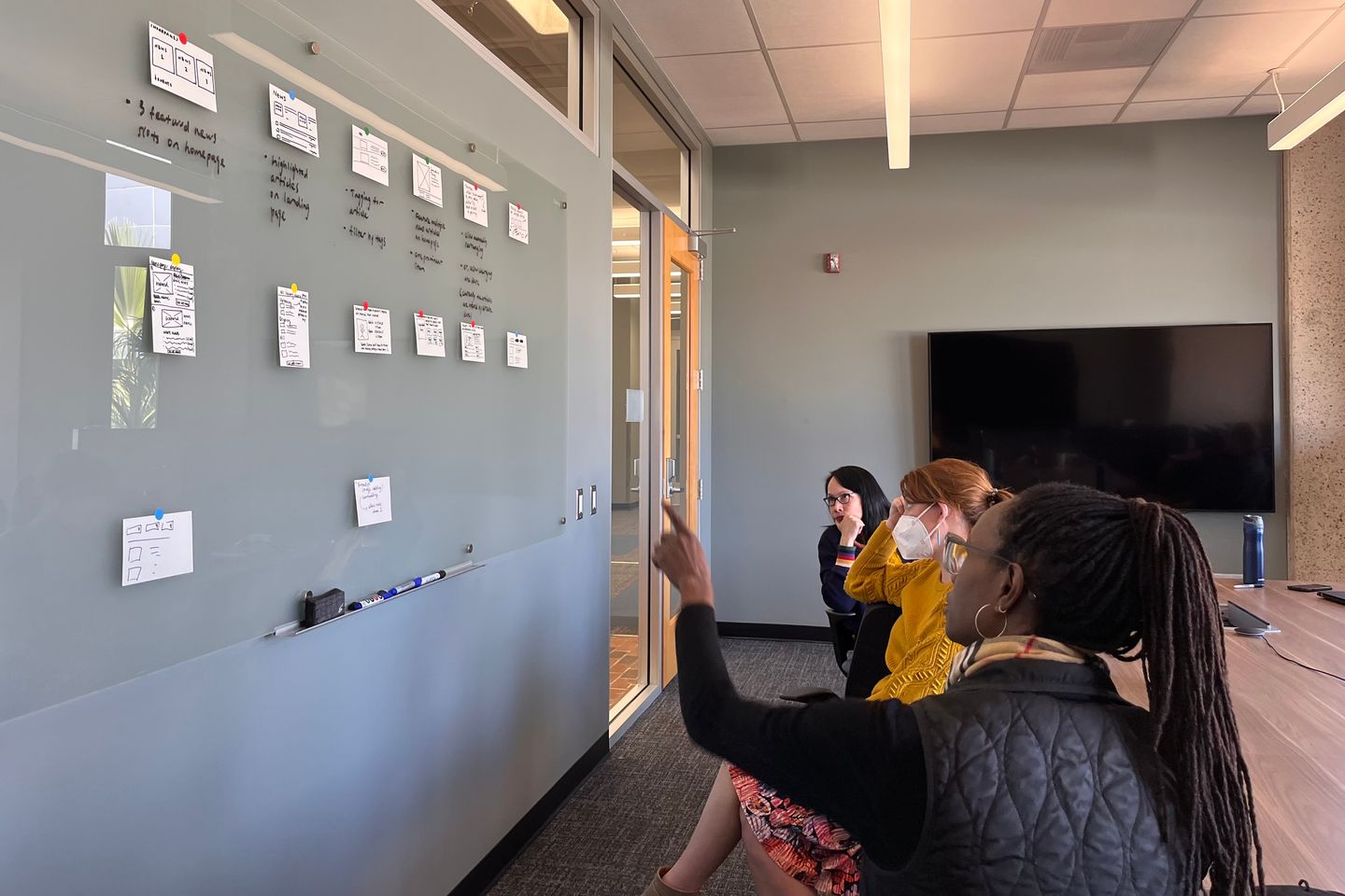 Three people sitting in front of a whiteboard with one women pointing at the whiteboard. Sketches on index cards are taped to the whiteboard.