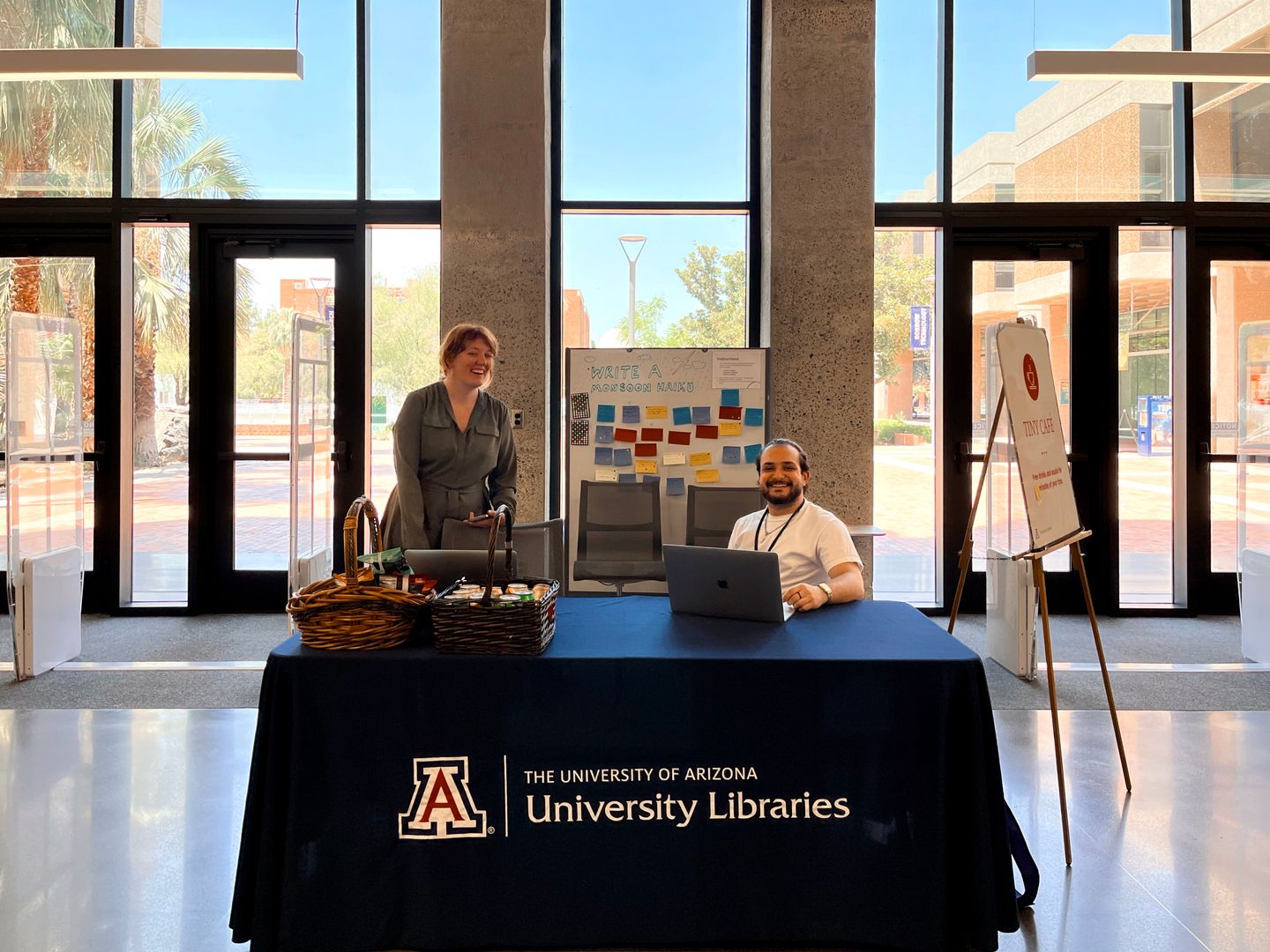 In a bright public space, two people behind a table labeled University Libraries with baskets of snacks and laptops on top of the table
